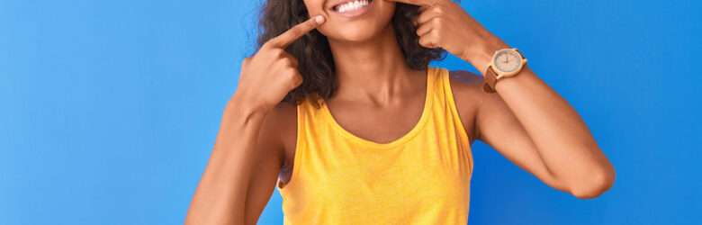 Brown woman against a blue wall in a yellow tank top smiles and points to her teeth after root canal therapy in Arvada, CO
